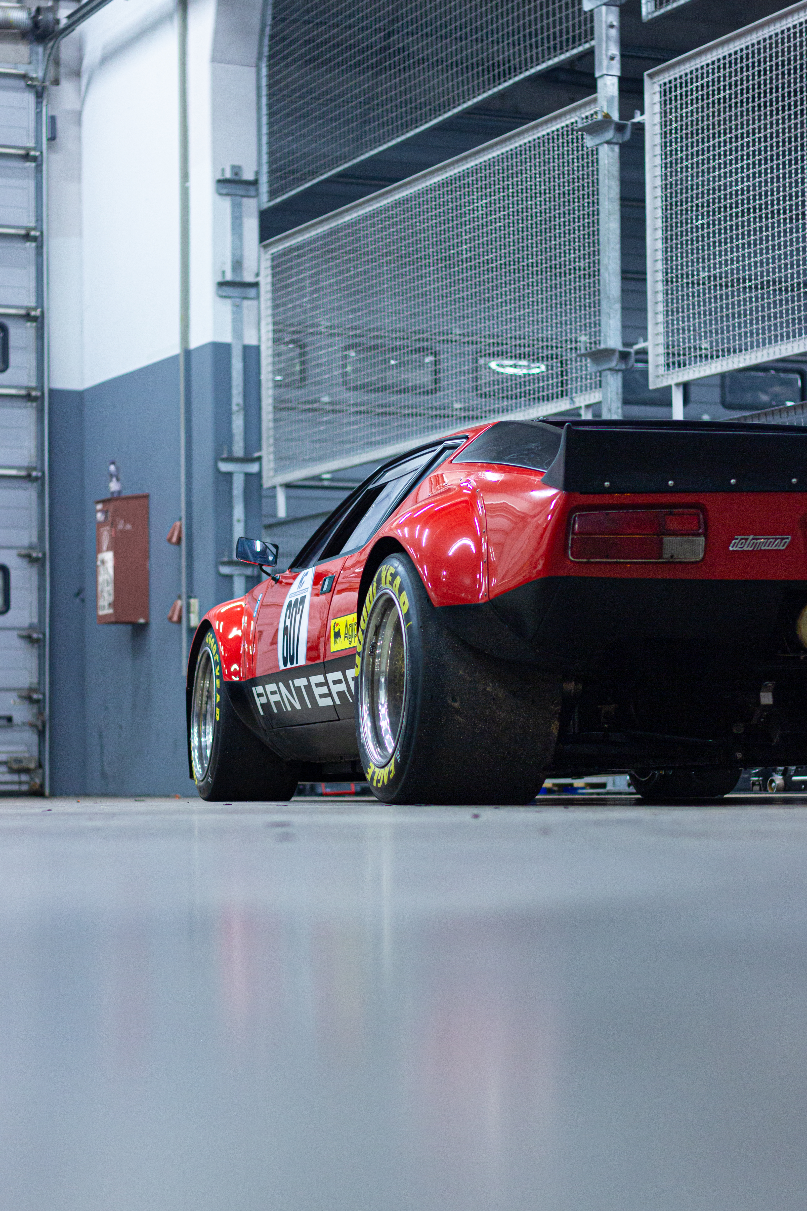 Roter De Tomaso Pantera Oldtimer in einer Box am Nürburgring, direkt an der Boxengasse aufgenommen. Der klassische Rennwagen steht mit breiten Slick-Reifen und markanter Karosserie bereit für den Einsatz auf der Rennstrecke.