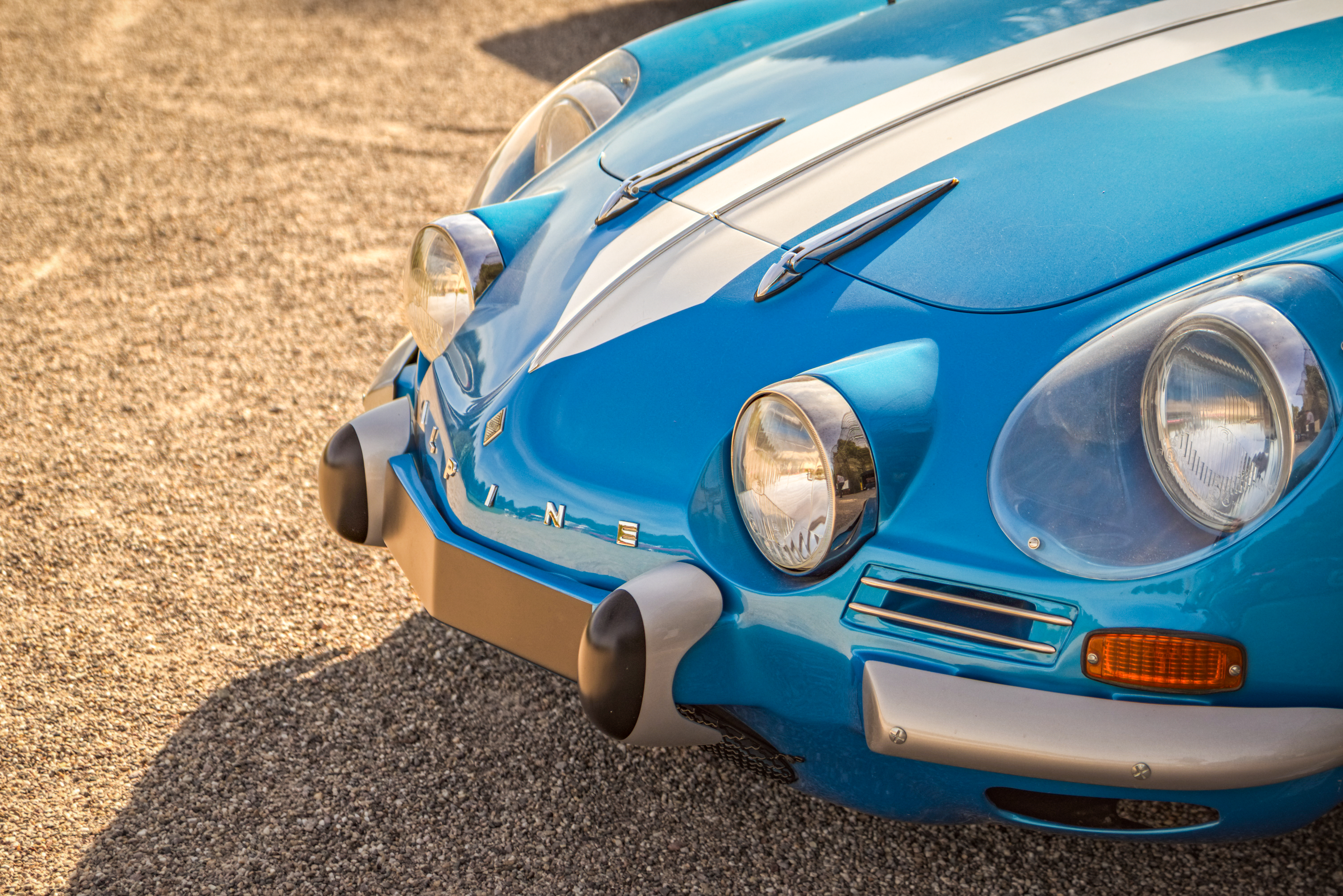 Fotografie eines blauen Oldtimers vom Typ Alpine A110 mit weißen Rallyestreifen im Motorsport Design – aufgenommen bei warmem Licht auf Schotter.