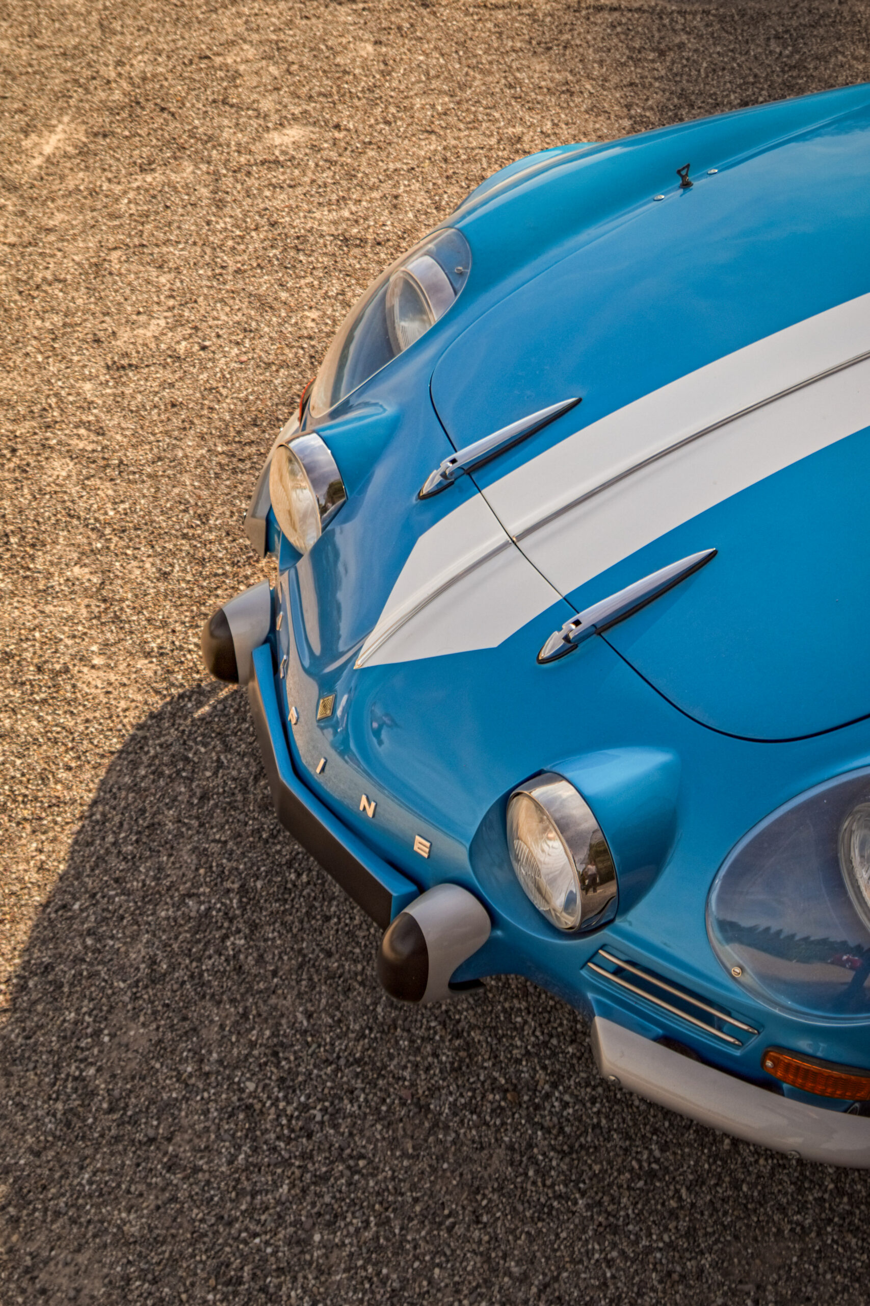 Fotografie eines blauen Oldtimers vom Typ Alpine A110 mit weißen Rallyestreifen im Motorsport Design – aufgenommen bei warmem Licht auf Schotter.